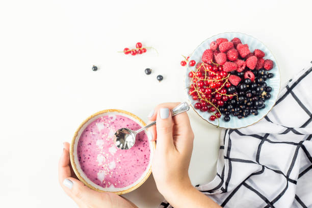 istock Female hands hold a berry smoothie in a bowl next to a plate with raspberries and black and red currants on a white table with a black and white checkered napkin. Healthy food concept. Top view 983995708