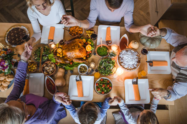Above view of extended family saying grace before Thanksgiving dinner. High angle view of extended family holding hands and praying before Thanksgiving dinner at dining table. saying grace stock pictures, royalty-free photos & images