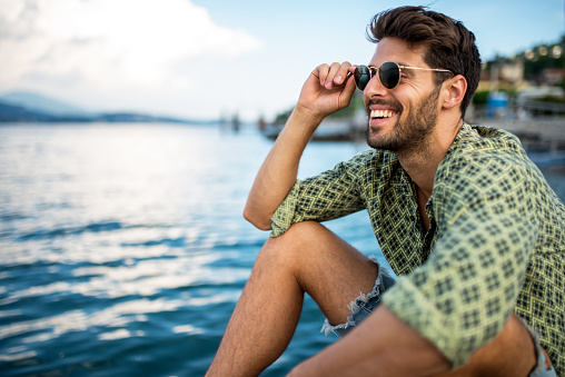 Handsome smiling man wearing sunglasses and looking away. He is sitting by the sea
