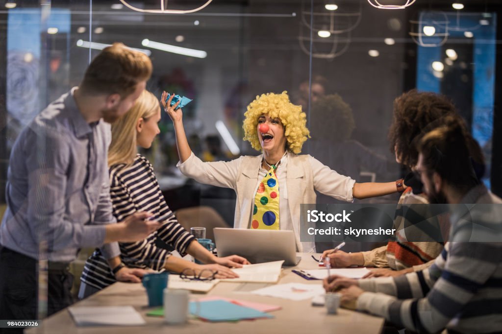 Playful businesswoman making a clown of herself on a meeting in the office. Young businesswoman having fun while pretending to be a clown on a meeting in the office. The view is through glass. Humor Stock Photo