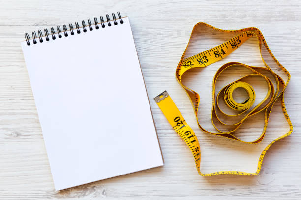 istock Measuring tape with notepad on a  white wooden surface, from above. Flat lay, top view. Copy space. Dieting concept. 984033612