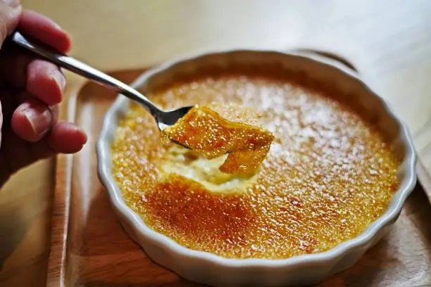 A hand of woman cracked through a crunchy top layer of a Creme brulee, burnt cream, crema catalana or Trinity cream, a rich and velvety custard base topped with a contrasting layer of hard caramel. Horizontal.
