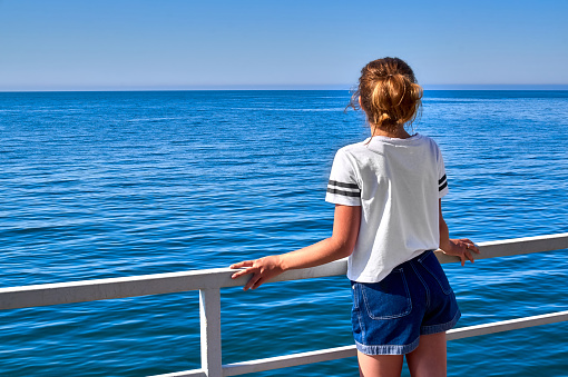 Young girl is standing on summer terrace with blue sea view. White railing.