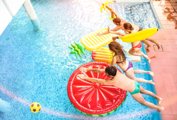 top view of active friends jumping at swimming pool party - vacation concept with happy guys and girls having fun in summer day at luxury resort - dynamic young people on warm bright sunshine filter - inflatable child jumping leisure games imagens e fotografias de stock