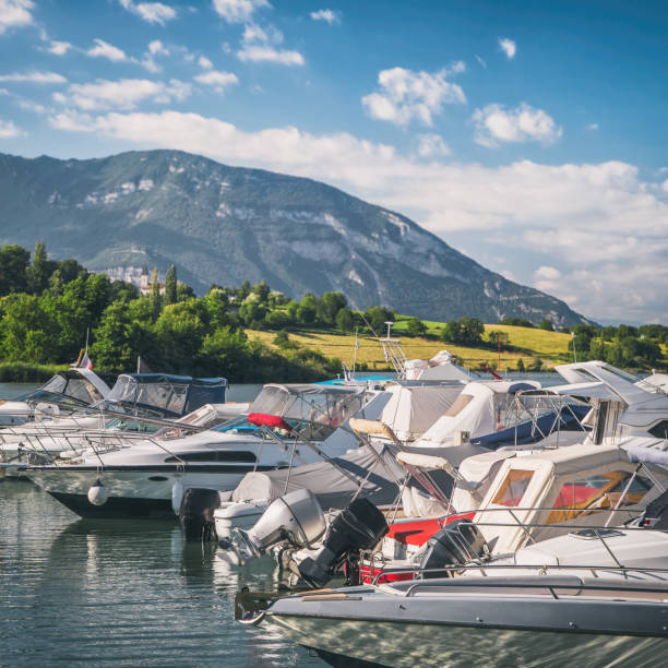 Square format color photography of small beautiful marina, harbor of Massignieu de Rives on Rhone river deviation, also named Lac du Lit Au Roi lake. There are several recreational motorized boats on water, with small French church steeple in meadow in middle of trees in background, blurred due to selective focus on nautical vessel in foreground, and famous mountain peak of Grand Colombier mountain away in background. This image was taken during a sunny summer day in European Alps, Bugey mountains in Ain department, Auvergne-Rhone-Alpes region in France (Europe).