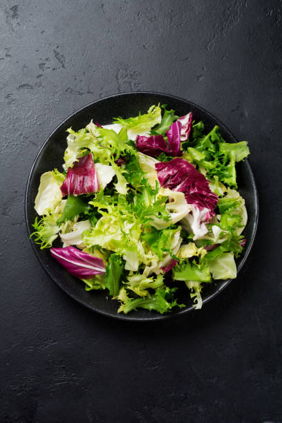 istock Fresh leaves of different salads in ceramic plate on black stone background. Selective focus. Top view. Copy space. 984286368