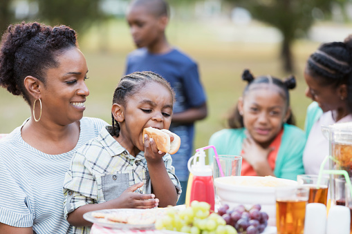 Two African-American adult sisters in their 30s with their three children at a backyard cookout. The children are mixed ages, 3 through 10 years old. The focus is on the mother and 3 year old boy biting into a hot dog.