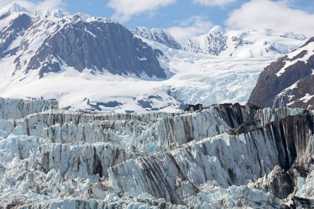 The Knik River and its Knik Glacier or Matanuska Glacier are starting to thaw and break apart.  The ice is a blue color, with the Alaska Mountain Range is the source of the river flow.  Ice, water and dirt are seen in the thawing ice and snow.