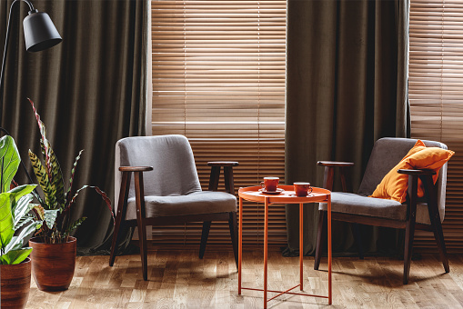 Mesa de centro sillones vintage, naranja con dos tazas, plantas de pie junto a la ventana con cortinas y persianas en el interior de una sala de estar photo