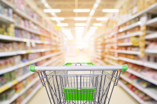 Supermarket aisle product shelves interior blur background with empty shopping cart