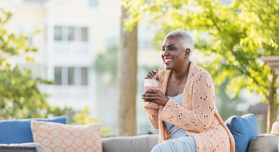 A senior African-American woman in her 60s relaxing outdoors on her patio, sipping a cold, refreshing beverage through a straw.