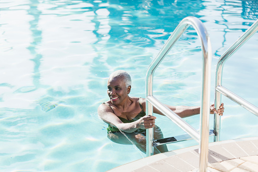 A senior African-American woman in her 60s in a swimming pool, smiling as she climbs out using the ladder.