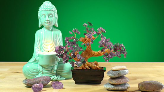 Calming zen interiors table setting with buddha statue holding burning candle, crystals, rocks against green background.