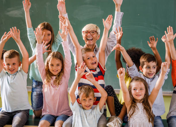 istock Happy teacher and elementary students with raised arms in the classroom. 984735236