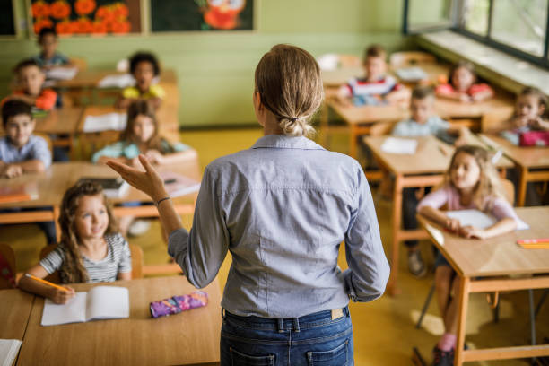 Rear view of a female elementary teacher giving a lesson in the classroom. Back view of a female teacher teaching large group of elementary students in the classroom. elementary school building photos stock pictures, royalty-free photos & images