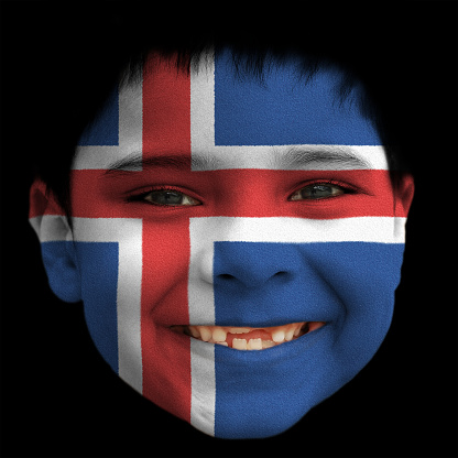 A lovely cheerful six year old boy smiling with broken incisors and half grown incisor teeth with Iceland  flag painted on his face.  Black background. Black hair falling on his forehead. Black eyes. white teeth. Face along with lips and ears is also painted