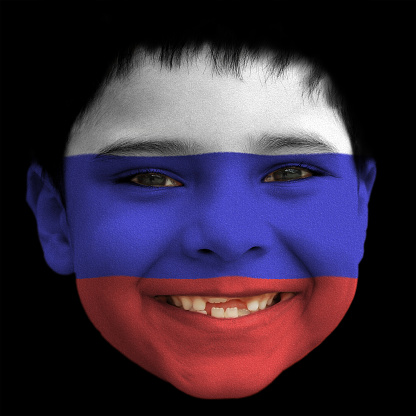 A lovely cheerful six year old boy smiling with broken incisors and half grown incisor teeth with Russia / Russian flag painted on his face.  Black background. Black hair falling on his forehead. Black eyes. white teeth. Face along with lips and ears is also painted