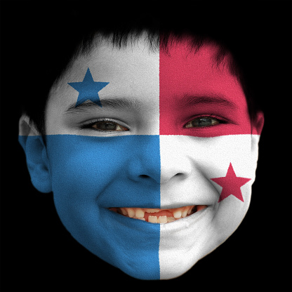 A lovely cheerful six year old boy smiling with broken incisors and half grown incisor teeth with Panama \n flag painted on his face.  Black background. Black hair falling on his forehead. Black eyes. white teeth. Face along with lips and ears is also painted
