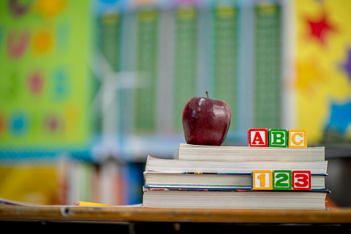 A stack of textbooks are sitting on a desk. An apple and toy blocks are on the books.