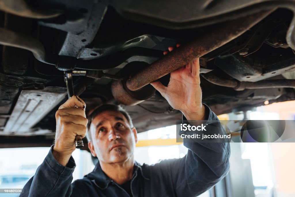 Mechanic working under a lifted car Man tightening a silencer pipe with wrench spanner in auto garage. Mechanic working under a lifted car in repair shop. Car Stock Photo