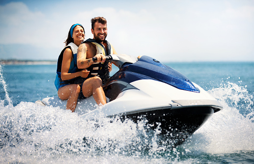 Closeup side view of a young couple riding a jet ski on a sunny summer day at open sea. The guy is driving and the girls is sitting behind.