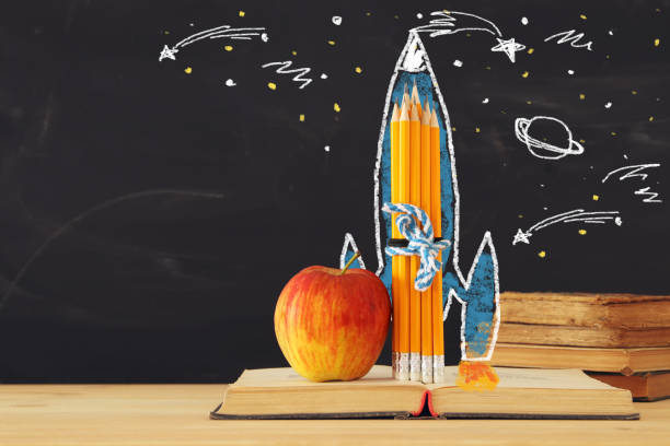 istock Back to school concept. rocket sketch and pencils over open book in front of classroom blackboard. 984846942
