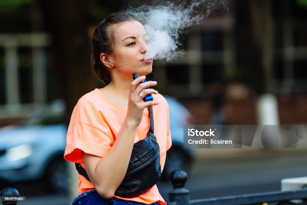 Pretty young hipster woman vape ecig, vaping device at the sunset. Toned image Pretty young girl vape popular ecig gadget,vaping device.Happy brunette vaper girl with e-cig.Portrait of smoker female model with electronic cigarette vaporizer.Ejuice vaping with fruit flavor liquid Electronic Cigarette Stock Photo