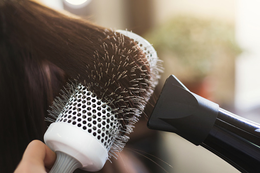 Hairdresser drying woman's hair with hair dryer and round brush in beauty salon, closeup