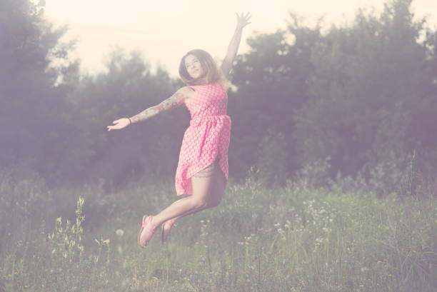 A joyful woman wearing a yellow dress is captured leaping gracefully amidst blooming trees in a sunlit urban park.