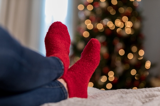 An unrecognizable woman stretches out her legs on a sofa during Christmastime. She is wearing fuzzy red socks.
