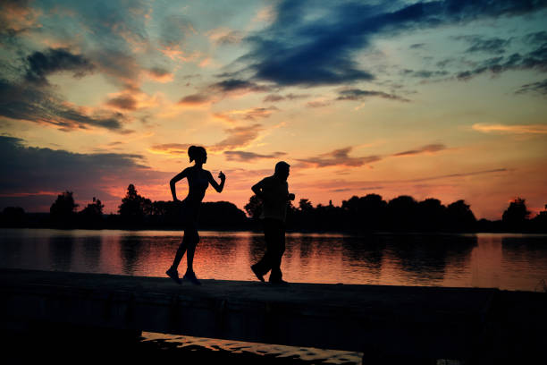 Dark silhouettes of scamper runners while sunset near lake. Slim young girl, senior man. Fitness, exercises. Fit, strong bodies, healthy lifestyle. Outdoors workout on fresh air.