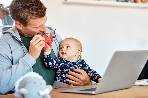 Father and baby boy playing with elephant toy with laptop on table. Mid adult man is spending leisure time with son at home. They are wearing casuals.