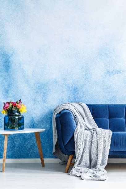 istock Comfortable dark blue settee with gray blanket and small side table with fresh flowers in a vase against blue and white ombre wall in a living room interior. Copy space background. Real photo. 985113930