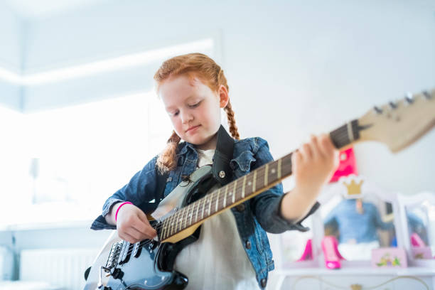 Girl playing electric guitar in here room at home. Young girl with long red hair playing rock music on electric guitar in her girlish room. Young female is practising playing electric guitar in her bedroom. Princess mirror is behind the girl. role reversal stock pictures, royalty-free photos & images