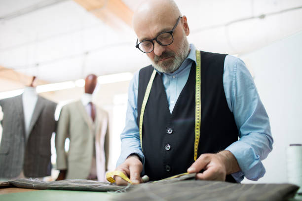 istock Serious concentrated bald skilled couturier with beard sewing menswear working with suit fabric and measuring it with soft tape in own workshop 985124580