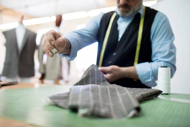 istock Close-up of busy senior tailor with thimble on finger stitching fabric for mens suit while working at table in own atelier 985124640