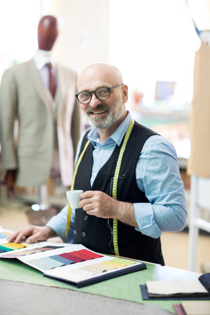istock Cheerful excited handsome bald senior bearded tailor with measuring tape on neck drinking espresso from small cup while choosing fabric from catalog with samples in sewing studio 985124762