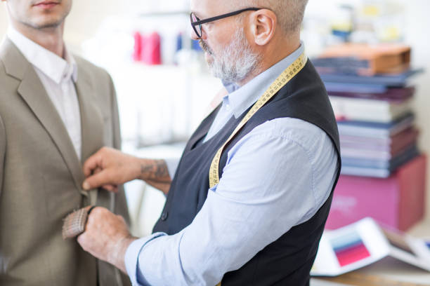 istock Close-up of content skilled senior bearded male tailor adjusting suit jacket while thinking of details for formal suit during fitting in atelier 985125064