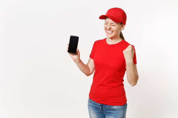 Delivery woman in red uniform isolated on white background. Female in cap, t-shirt, jeans working as courier or dealer showing on camera mobile phone with blank empty screen. Copy space advertisement. Delivery woman in red uniform isolated on white background. Female in cap, t-shirt, jeans working as courier or dealer showing on camera mobile phone with blank empty screen. Copy space advertisement niñas stock pictures, royalty-free photos & images