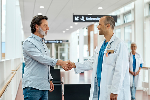 Male doctor smiling while shaking hands with patient in waiting room. Mature male is greeting doctor in medical clinic. They are standing in hospital.