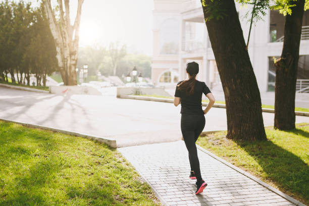 istock Back view Young athletic brunette girl in black uniform and cap training, doing sport exercises and running, looking straight on path in city park outdoors. Fitness, healthy lifestyle concept. 985132408