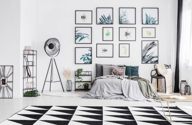 istock King-size bed with many pillows and grey sheets standing in white bedroom interior with big studio lamp, black and white carpet and plastic chair with knit blanket 985150558