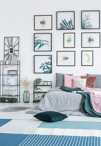 Green cushion placed on the floor with blue and white carpet in bright bedroom interior with double bed. two metal racks with plants and decor and simple gallery posters hanging on the wall