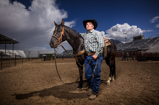 Dramatic portrait style shot of a man in western wear standing beside a horse taken at a rodeo arena, using artificial light.
