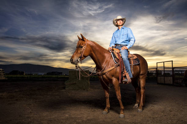 Portrait of a young man in western wear and a cowboy hat, sitting on horseback in a paddock on a ranch in Utah. Shot with artificial light against a dramatically setting sun.
