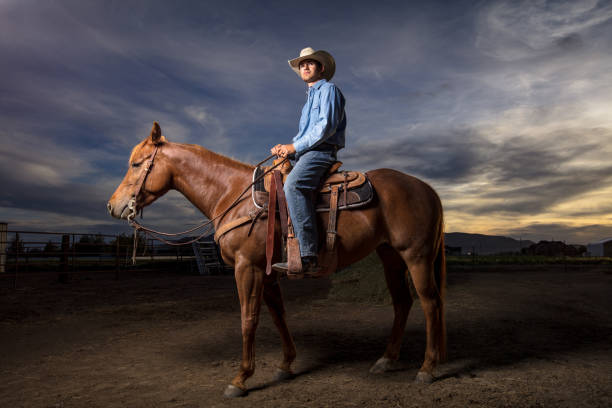 Portrait of a young man in western wear and a cowboy hat, sitting on horseback in a paddock on a ranch in Utah. Shot with artificial light against a dramatically setting sun.