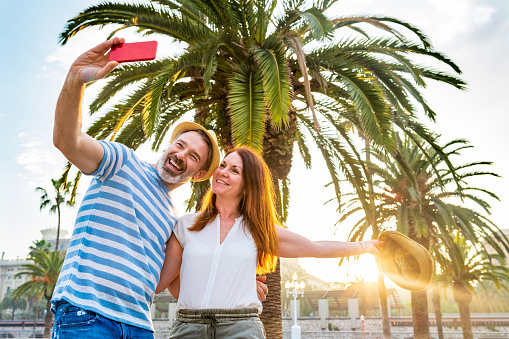 Adult couple dating and having fun in Barcelona at sunset. Man and woman together on a summer sunny day in Barcelona, taking a selfie with palm trees and sun on background. Love and lifestyle concepts