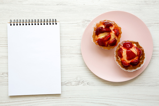 Strawberry vanilla cream cheese tarts on pink plate with notebook, top view. From above, overhead.