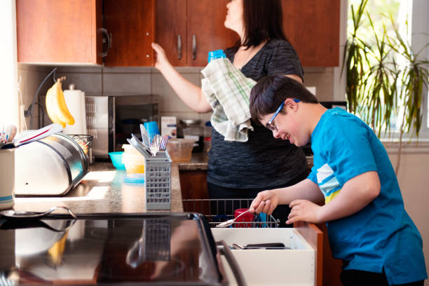 Mother helping her son of 12 years old with Autism and Down Syndrome in daily lives emptying the dishwasher stock photo