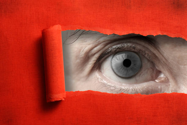 istock Gray eyes looking through from a ripped hole in red paper 985442454